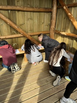 Learners in the outdoors, brainstorming their personal stories together in pairs. One learner is holding a small rock in their hand. A teacher is talking with one of the learners. A learner holds up a pen together with a cone they found while brainstorming. Photo: Marte Maurabakken/Climate Creativity. 