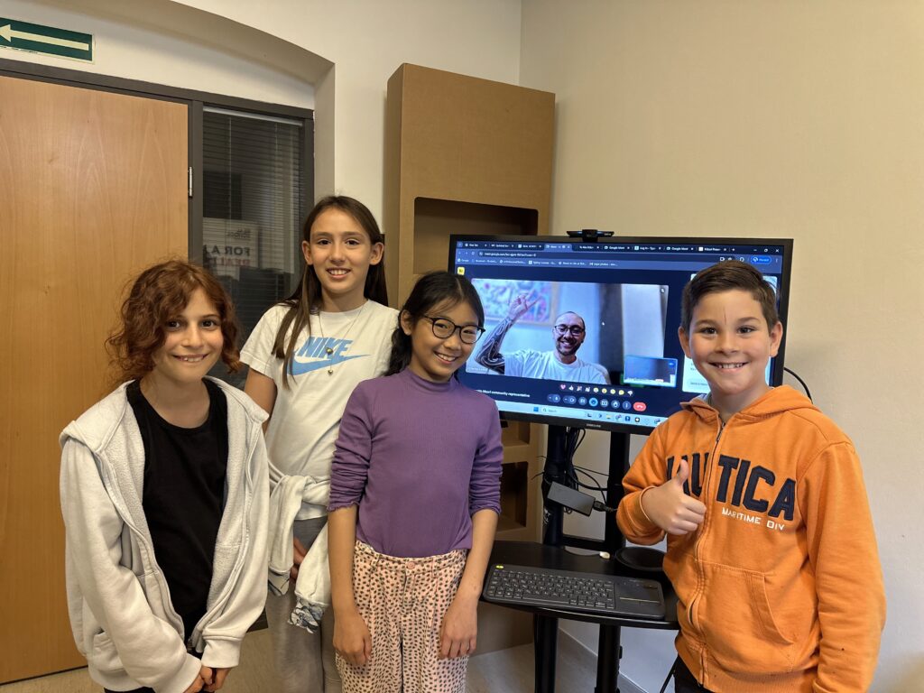 4 children are standing around a screen showing a video conversation with an adult. Everyone are smiling.